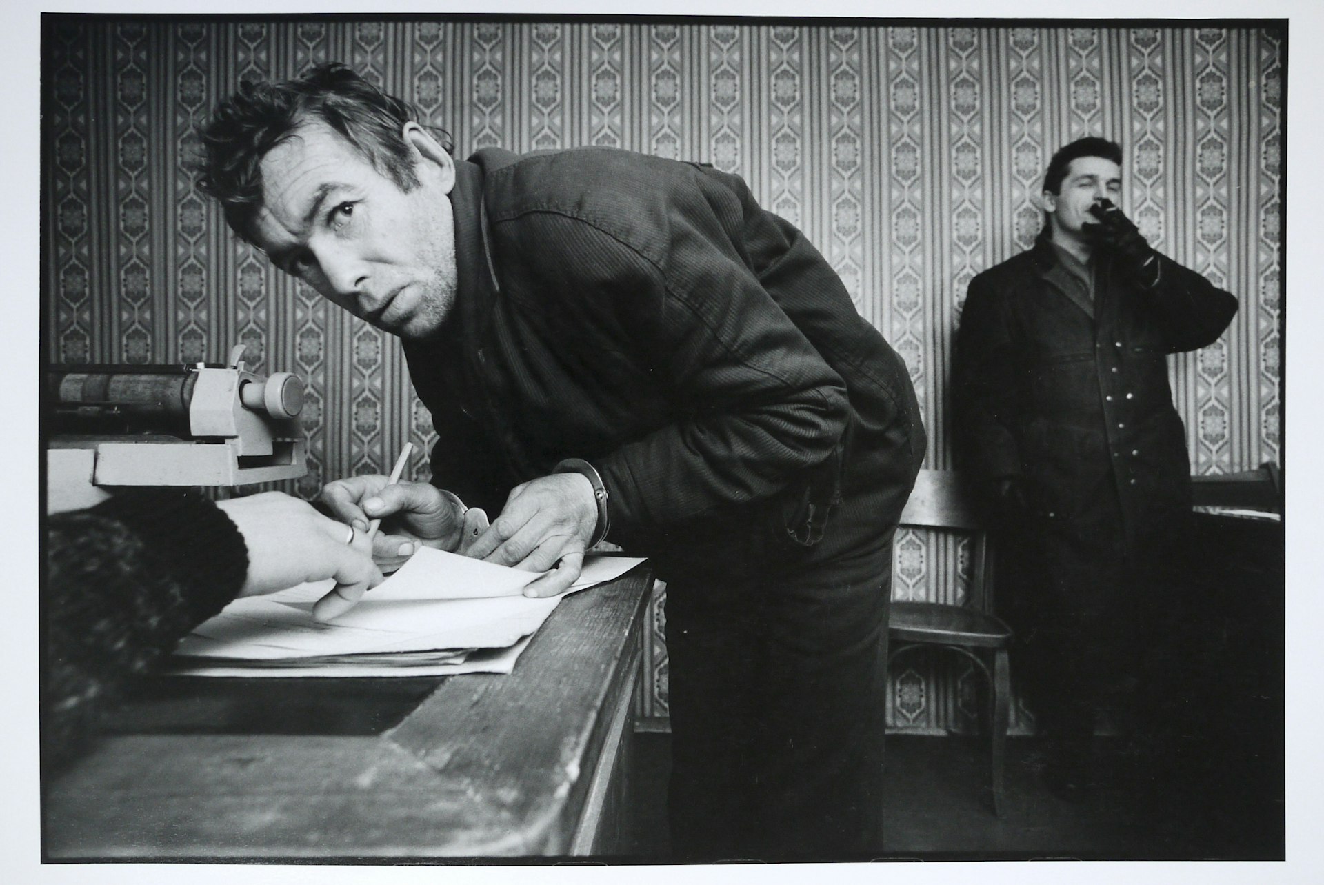 A petty thief signing his confession at the local police station. The arresting officer stands in the background, December 1992.