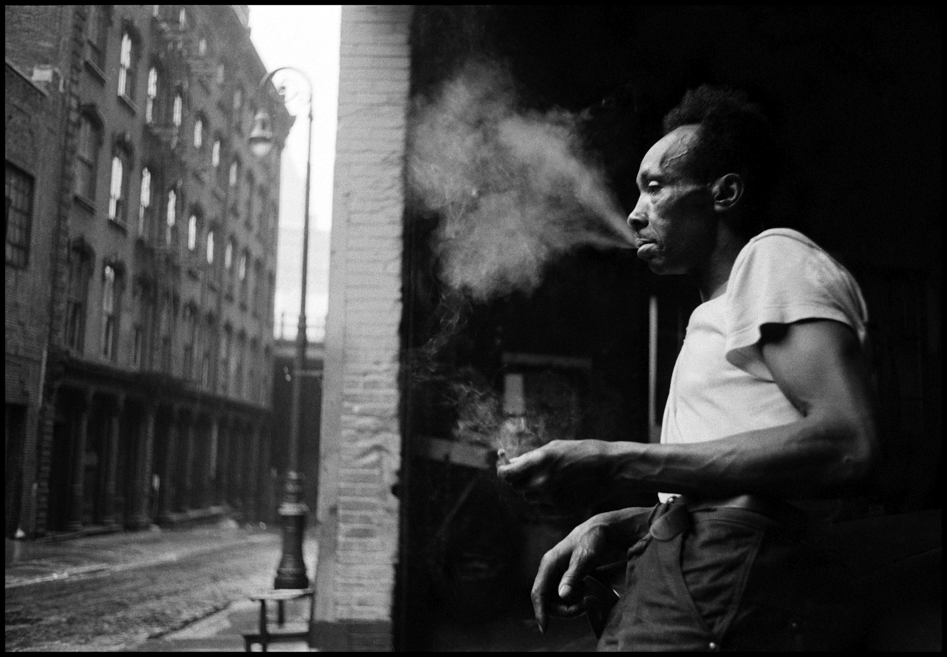 USA. New York City. 1955. Man smoking in the streets under the Brooklyn Bridge. Photo by Erich Hartmann.