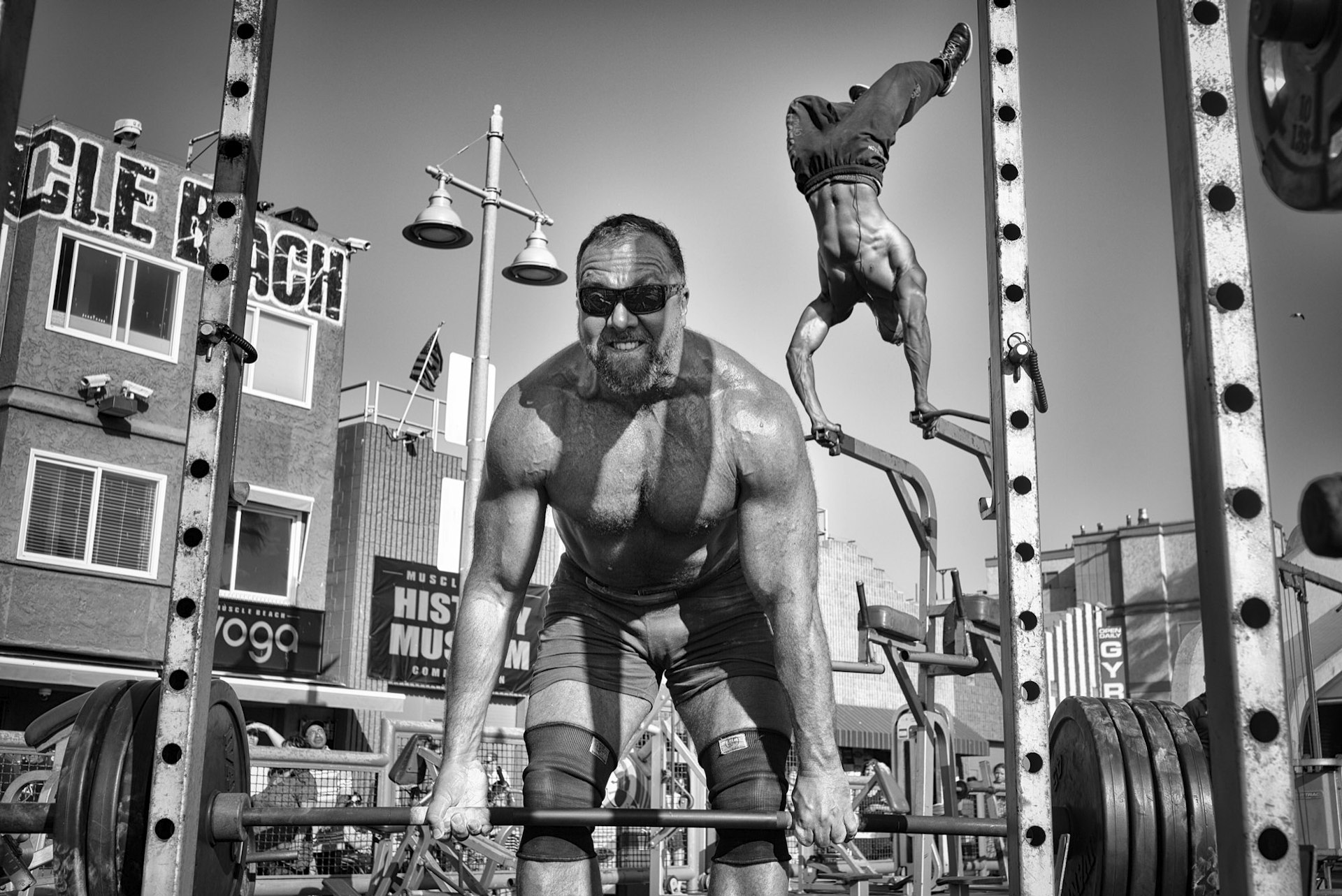 A weightlifter raises a barbell loaded with heavy plates while Ike Catcher performs an aerial handstand at the world-famous Muscle Beach Gym. © Dotan Saguy