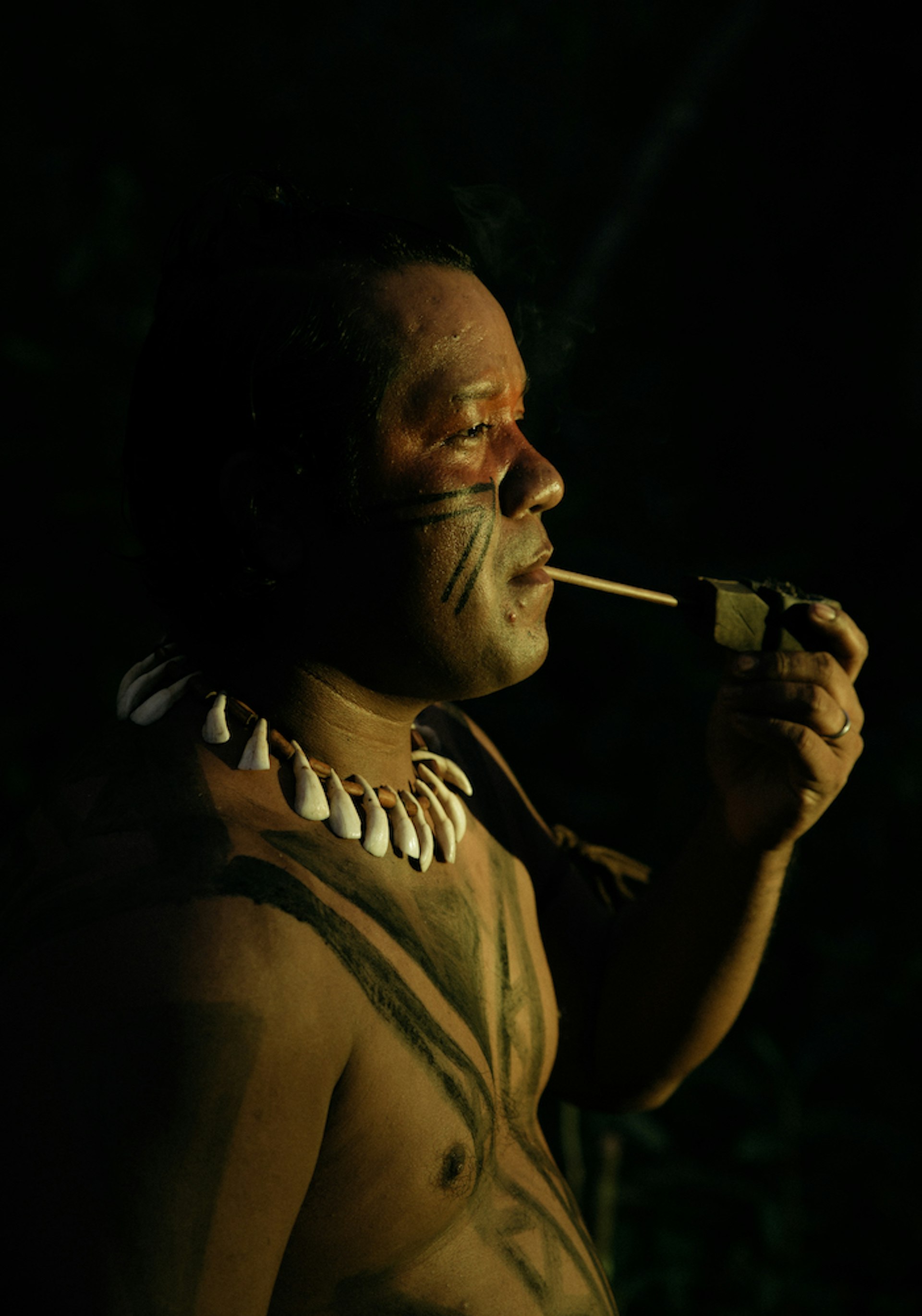 an indigenous brazilian man smokes a carved pipe. He is shirtless with his chest adorned in body paint and wears a necklace.