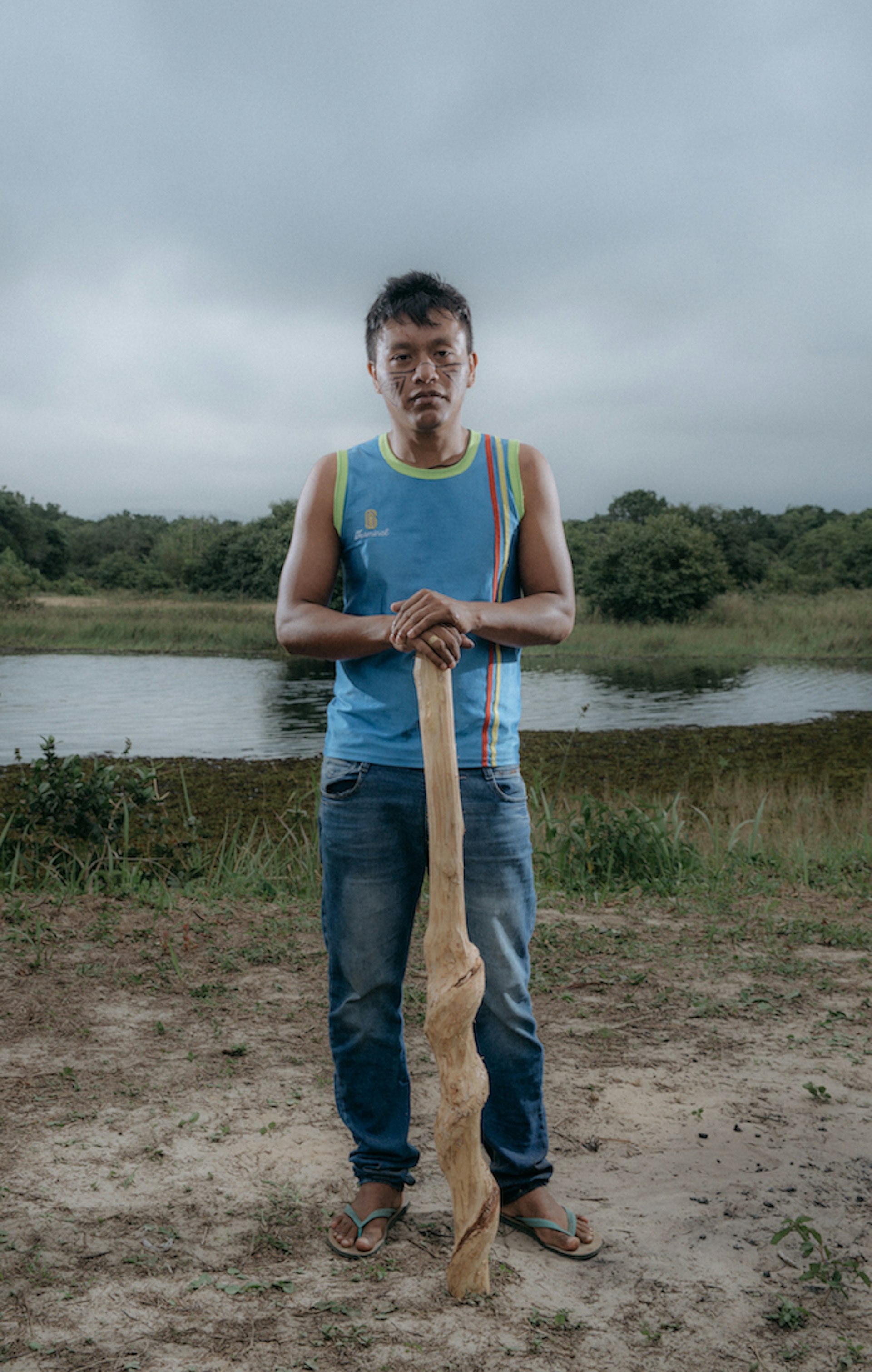 An indigenous Brazilian man standing in front of a small lake. He wears modern clothing and rests his hands on top of a large, carved wooden club.